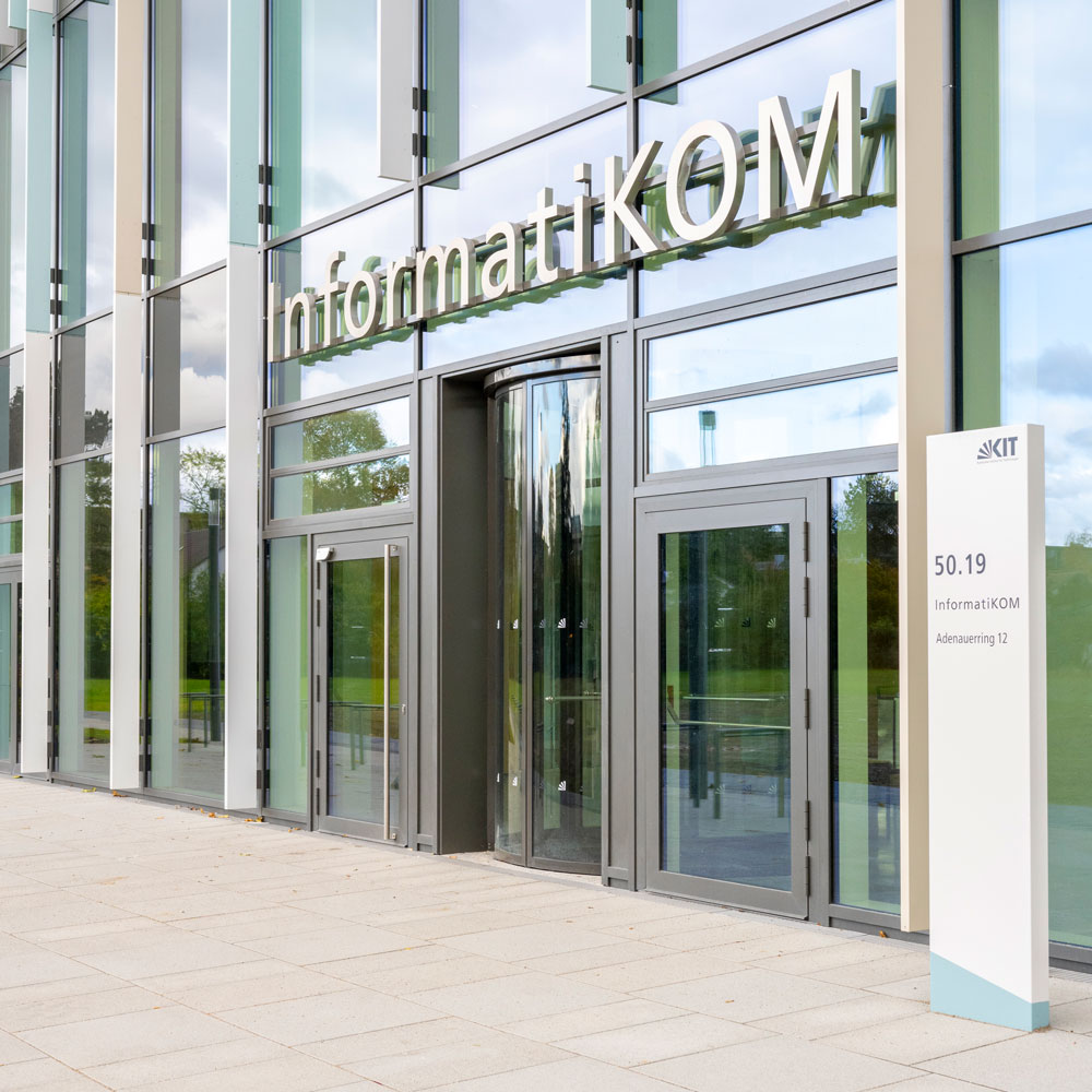 Außenansicht eines modernen Gebäudes mit Glasfront, Beschilderung "InformatikKOM" und Eingangsbereich.